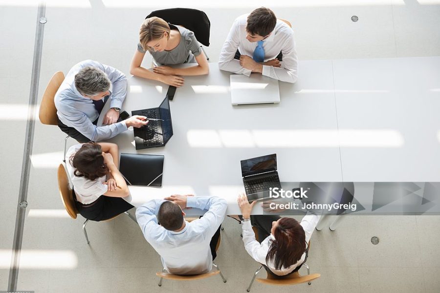 Business People In Meeting. A group of six men and women (three man and three woman) sitting around an oval white table having a business meeting. They all are dressed in business causal, and have their laptops out, only two are being used. They are sitting in simple wooden chairs.